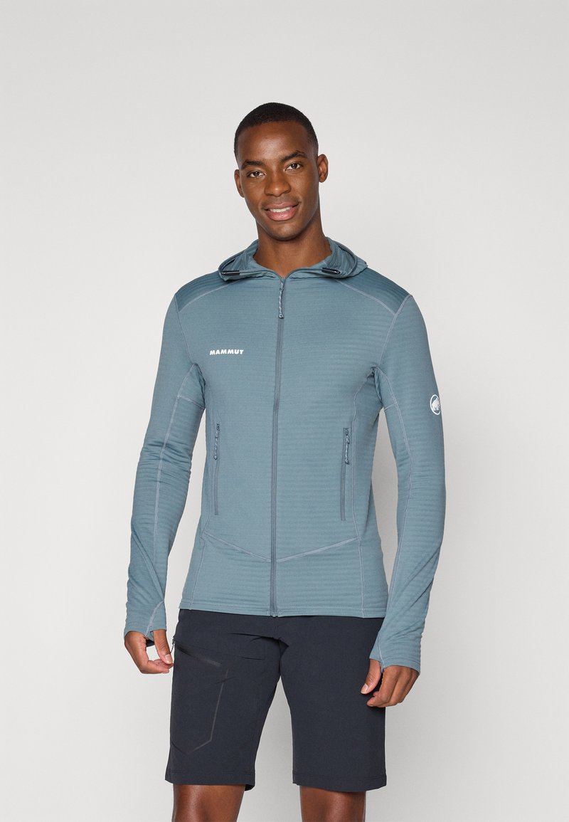 Leichte, kapuzenjacke aus blau-grauem strukturiertem Stoff mit figurbetontem Design, langen Ärmeln und mit Reißverschlüssen versehenen Taschen. Kombiniert mit dunklen Shorts.