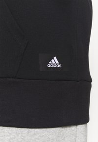 Gros plan sur l'ourlet d'un sweat-shirt Adidas noir avec un patch logo au-dessus de la ceinture côtelée, porté sur un tissu gris clair.