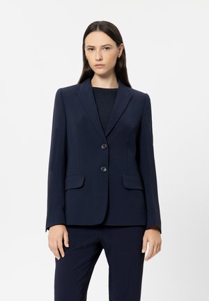 Frau mit langen dunklen Haaren, die einen taillierten marineblauen Blazer und passende Hose vor einem schlichten hellen Hintergrund trägt.