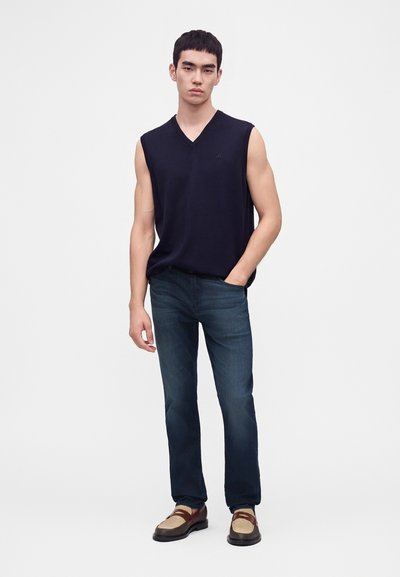 Jeune homme portant un haut noir sans manches en V, jeans bleus et mocassins marron bicolores, debout avec une main dans la poche.
