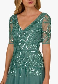 Kobieta ubrana w turkusową sukienkę z cekinowymi wzorami liści i przezroczystymi półdługimi rękawami, z dekoltem w kształcie litery V i dopasowaną talią.