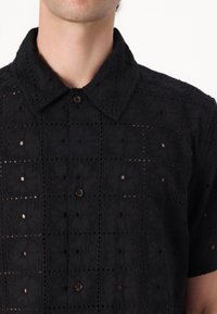 Chemise noire à manches courtes avec un motif brodé et des découpes géométriques, un col pointu et une fermeture à boutons. Tissu texturé.