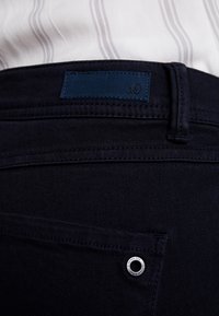 Tmavě modré džíny s hladkým povrchem, vybavené koženou nášivkou, kovovým očkem a viditelným švem na zadní kapse.