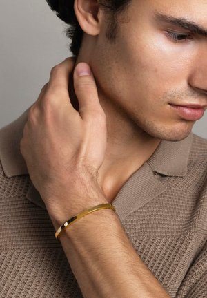 Gold bangle bracelet worn on wrist, smooth surface with subtle shine, set against a textured beige sweater. Skin tone is light with fine hair.