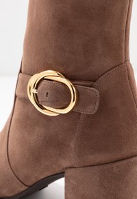 Bottine en suède marron avec un bout arrondi et une boucle en or décorative sur une sangle. Dispose d'un talon épais et d'une texture lisse.
