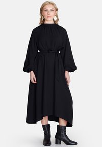 Rochie midi neagră cu mâneci lungi tip balon, talie strânsă cu un cordon și tiv asimetric. Asortată cu cizme negre până la gleznă. Textură de material neted.