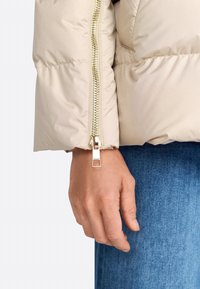 Beige pufferjack met een glanzende gouden rits en kenmerkende hardwaredetails. Zachte, gekwilt textuur; close-up van de mouw en hand.