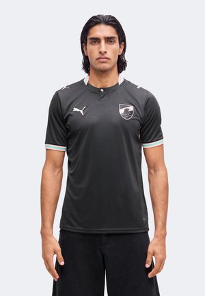 Homme aux cheveux foncés mi-longs, portant un t-shirt noir Puma avec un blason et une bordure rayée sur les manches, debout devant un fond uni.