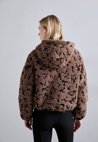Brown fleece jacket with a black abstract pattern, featuring a hood and elastic hem. Textured material provides warmth and softness.
