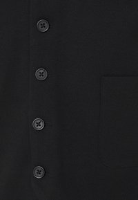 Gros plan sur un tissu noir avec quatre boutons noirs alignés verticalement et le contour d'une poche sur le côté droit.