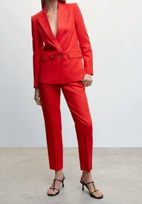 Una donna in un completo rosso su misura si erge con sicurezza, abbinato a tacchi a listini neri, su uno sfondo neutro.