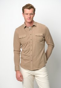 Camicia a maniche lunghe color marrone chiaro realizzata in tessuto morbido, con apertura a bottoni, due tasche sul petto e bottoni a pressione argentati.