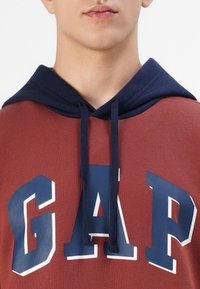 Jeune personne portant un sweat à capuche couleur rouille avec une capuche bleu marine et "GAP" imprimé en grandes lettres bleu marine sur la poitrine.