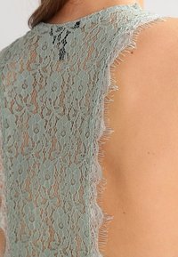 Lace topp i ljusgrön med blommönster och fransade kanter. Genomskinlig textur avslöjar huden under. Närbild av bakdesignen.