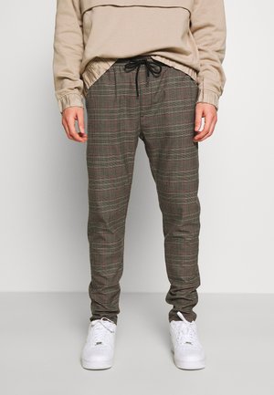 Mann trägt beige Sweatshirt, graue karierte Jogginghosen mit schwarzem Kordelzug und weiße Sneakers, steht vor weißem Hintergrund.