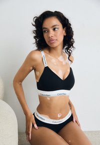 Kvinne med vitiligo iført svart og hvit Calvin Klein undertøy, sittende mot en nøytral bakgrunn og ser til siden.