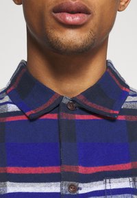 Gros plan sur la partie inférieure du visage et le cou d'une personne portant une chemise à carreaux bleue, rouge, blanche et noire avec col visible.