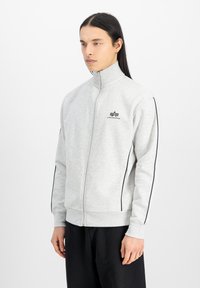 Graue Zip-Jacke mit hohem Kragen, schwarzen Seitenstreifen und Logo auf der Brust. Verfügt über gerippte Bündchen und eine glatte, weiche Textur.