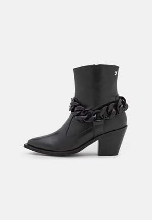 Bottine noire en cuir avec un bout pointu, un talon moyen épais et une grande chaîne noire décorative autour de la cheville.