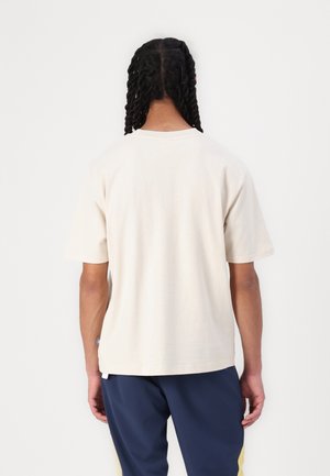 Kremfarvet t-shirt med korte ærmer, bomuldsstof, afslappet pasform, rund halsudskæring, båret med navybukser, synlig mærkat på kanten.