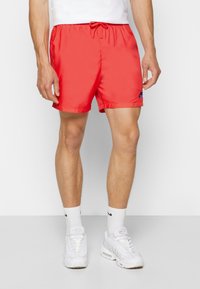 Muž oblečený v jasně červených Nike šortkách s modrým logem, bílých Nike ponožkách a bílých teniskách stojící na jednoduchém bílém pozadí.