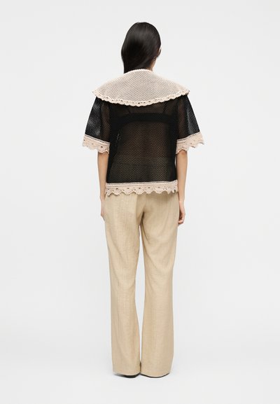 Γυναίκα με πλάτη, που φοράει μαύρο τοπ από δίχτυ με μπεζ πλεκτή διακόσμηση και φαρδιά μπεζ παντελόνια, σε λευκό φόντο.