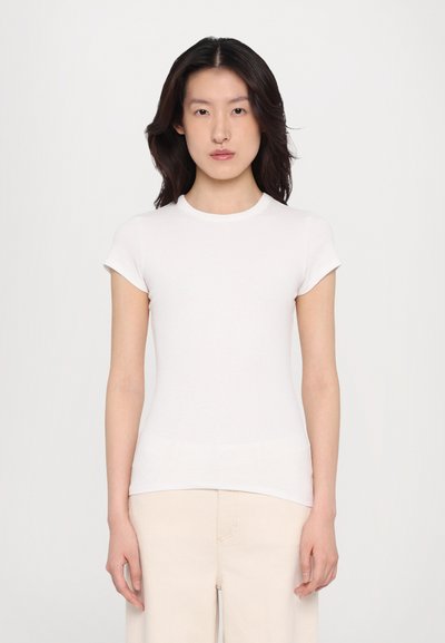 T-shirt blanc à manches courtes, ajusté, en tissu côtelé ; doté d'un col rond et d'épaules sans couture. Porté avec un pantalon de couleur claire.