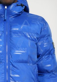 Chaqueta acolchada azul con acabado brillante, que incluye capucha, cierre con cremallera y un patrón de logo en toda la superficie para añadir textura y detalle de marca.