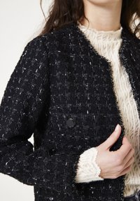 Zwarte tweed jas met een gestructureerd patroon, voorzien van een franjerand en een ronde zwarte knoop. Daaronder is een crèmekleurige gebreide trui zichtbaar.