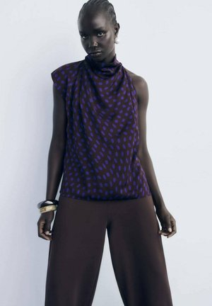 Top viola con pois blu, design drappeggiato e spalla asimmetrica. Abbinato a pantaloni larghi marroni e braccialetti chunky.