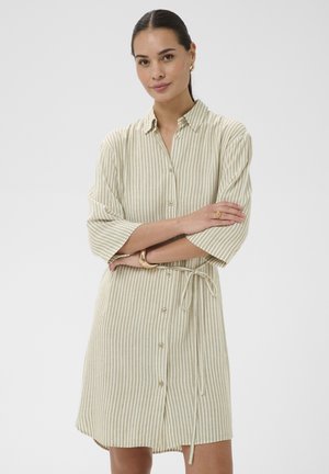 Žena s tmavými vlasmi v béžových a bielych vertikálne pruhovaných šatách na gombíky so štvrťrukávmi, sedia zlaté kruhové náušnice a náramky.