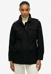 Chaqueta negra de estilo militar con cremallera frontal, dos bolsillos en el pecho y hombreras. Hecha de un tejido duradero, con un cuello clásico.