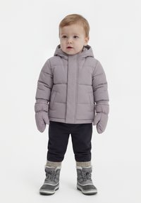 Bambino piccolo indossa una giacca imbottita grigia con cappuccio, muffole abbinate, pantaloni neri, calzini spessi e stivali invernali grigi in piedi su uno sfondo bianco.