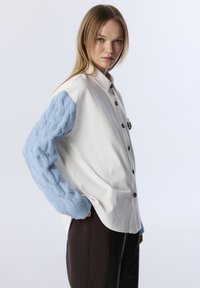Chemise boutonnée avec un corps crème et des manches en maille torsadée bleu clair. Elle présente un col classique et des boutons foncés, portée avec un pantalon foncé.