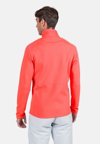 Ljus korallfärgad fleecejacka med fjärdedel dragkedja och långa ärmar, med hög krage och mjuk, elastisk textur. Lätt och figurnära design.