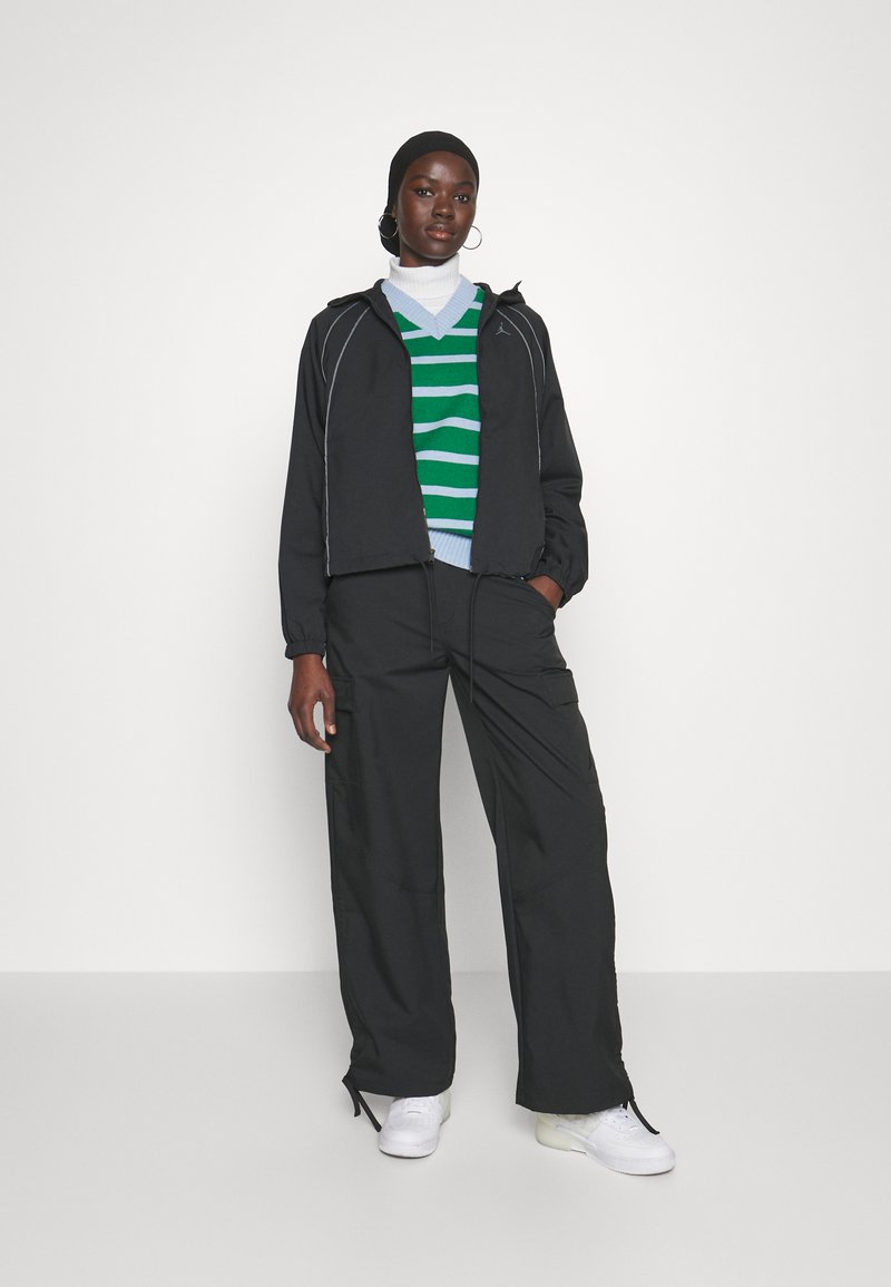 Chaqueta negra con cremallera y detalles en blanco, suéter a rayas verdes y blancas debajo, y pantalones cargo negros. Zapatillas blancas completan el atuendo.