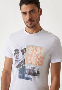 Biela bavlnená tričko s multikolorovou grafikou a nápisom "GUESS" a rôznymi textúrovanými obrazovými záplatami v odtieňoch modrej a oranžovej.