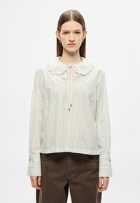 Biała bluzka z pionowymi czarnymi paskami, z dużym, zaokrąglonym kołnierzem, wiązanym detalem przy szyi oraz mankietami na guziki. Miękki materiał, luźny krój.