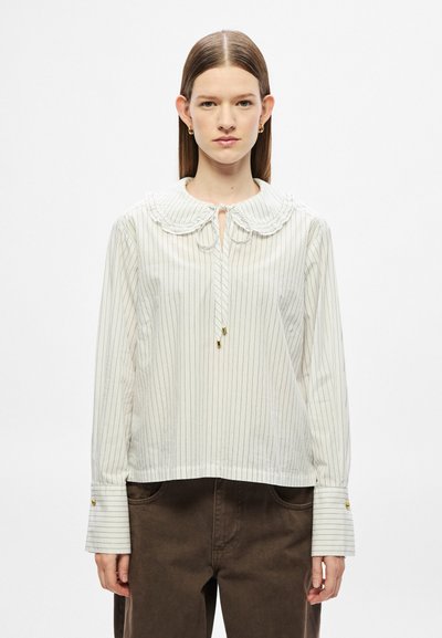 Blouse blanche à rayures noires verticales, avec un grand col rond, un détail noué au niveau du cou et des poignets boutonnés. Tissu doux, coupe ample.