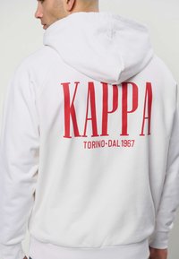 Mees, kes kannab valget kapuutsiga pusa, mille seljal on suur punane kiri "KAPPA TORINO DAL 1967", seisab veidi vasakule pöördunult ja kapuuts on alla pandud.