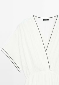 Robe blanche avec un décolleté en V et des manches courtes, présentant un passepoil noir le long des bords et un tissu texturé. Détail de taille élastique.