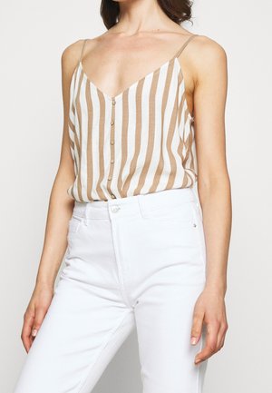 Vrouw draagt een beige en witte verticaal gestreepte mouwloze top met knopen, ingestopt in een witte broek met hoge taille.
