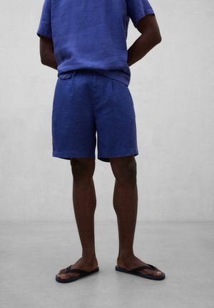 Mann iført blå kortermet skjorte, matchende blå shorts og svarte flip-flops som står mot grå bakgrunn.