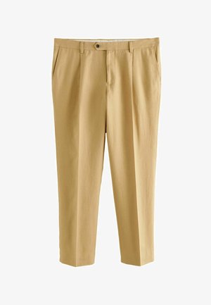 Man draagt beige broek met knoopsluiting en een zwarte gebreide top, arm ontspannen langs het lichaam tegen een neutrale achtergrond.