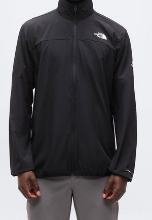 Osoba nosząca czarną kurtkę z zamkiem błyskawicznym z logo The North Face na piersi i lewym rękawie, w połączeniu z jasnoszarymi spodniami.