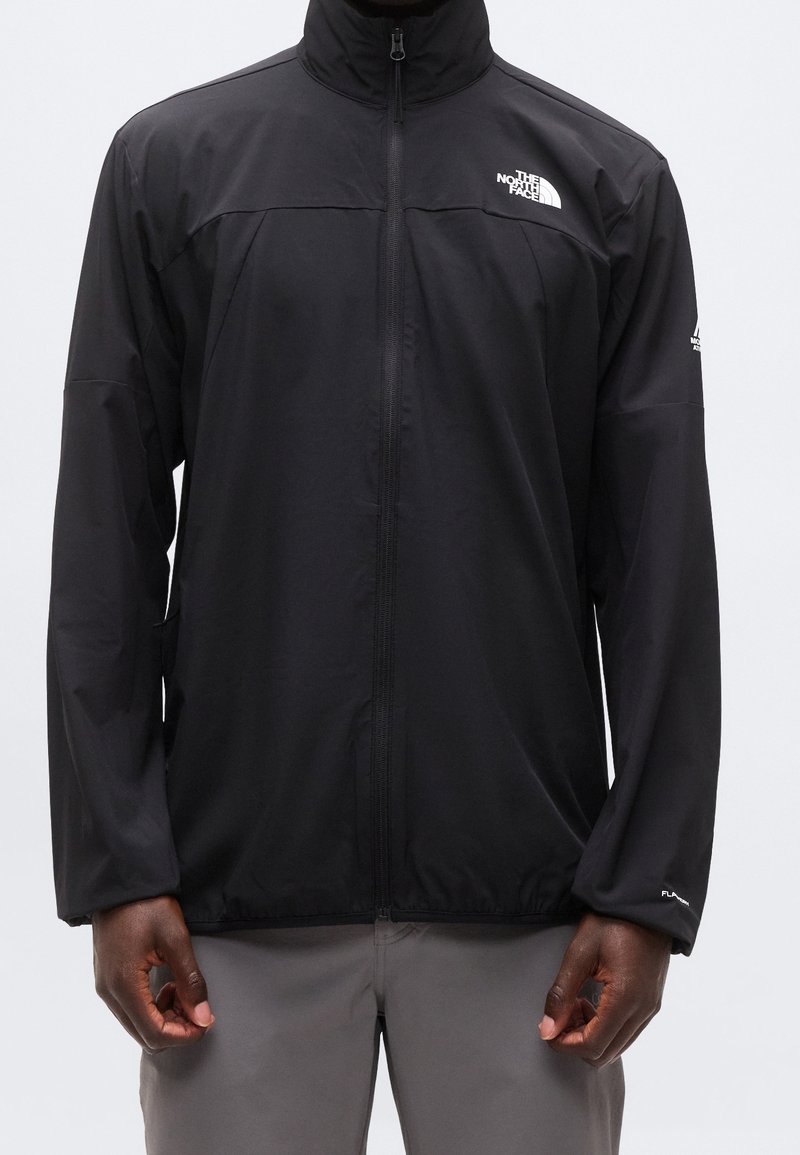 Personne portant une veste noire zippée avec le logo The North Face sur la poitrine et la manche gauche, associée à un pantalon gris clair.