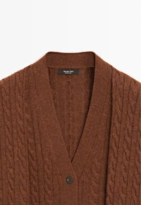 Hnědý pletený cardigan se V-výstřihem, se vzorem z copánkového pletení a jediným černým knoflíkem na zapínání, vyrobený z měkké tkaniny.