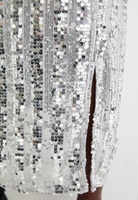 Robe en sequins argentés avec des lignes verticales, comportant une fente sur le côté. Le tissu est léger, avec une texture brillante et des accents réfléchissants.