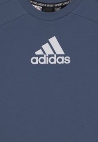 Sininen urheilupaita, joka on valmistettu sileästä kankaasta, ja jossa on valkoinen "adidas"-logo selkeästi rinnassa. Pyöreä kaula-aukko ja yksinkertainen muotoilu.
