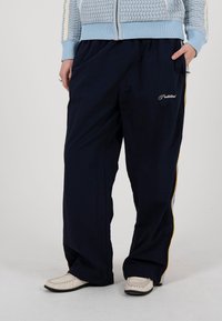 Žmogus dėvi tamsiai mėlynas plačias kelnes su geltonomis šoninėmis juostomis, šviesiai mėlyną megztą megztinį ir baltus loaferius, stovintis su rankomis kišenėse.